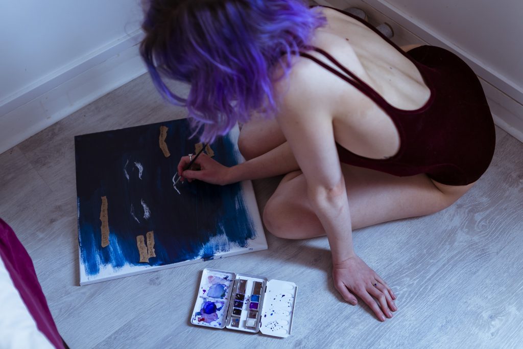 Un·e artiste est penché·e sur sa toile et peint. Iel a les cheveux violets et sa toile est dans les tons de bleus.