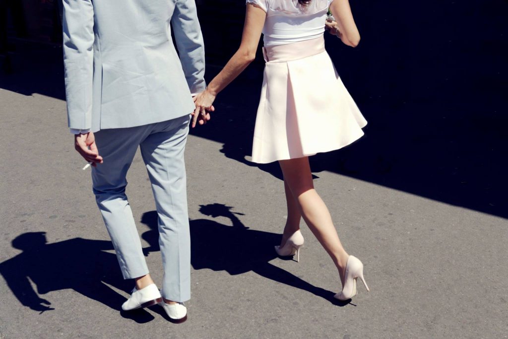 Deux personnes se tiennent la main en marchant dans la rue. Ils sont de dos. Le soleil leur tape dans le dos ce qui forme leurs ombres bien dessinées au sol. La personne de gauche porte un costume bleu ciel et celle de droite, une jupe rose pâle.