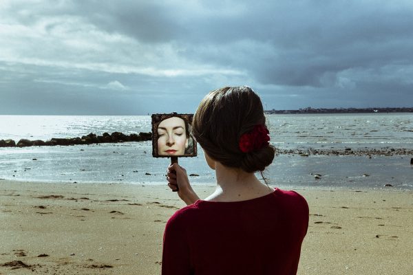 Une personne aux cheveux longs ramassés en chignon tenu par une fleur rouge se tient de dos, face à l'océan. Elle porte un pull de la même couleur que la fleur dans ses cheveux. Elle tient un miroir dans sa main, on peut donc apercevoir une partie de son visage dans le reflet. Elle a les yeux fermés et semble sereine.